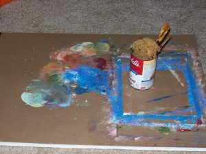 An impromptu cardboard palette, soup can water cup, paint brushes, and sponge